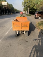 A person wearing a hard hat is driving an orange utility vehicle down an empty road lined with trees. The vehicle is loaded with tools, and a shadow of a bicycle appears on the road surface.