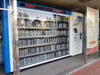 A vending machine filled with neatly arranged books is situated outdoors. There is a digital display above the machine showing text in a language that appears to be Chinese. Next to the machine is a panel with information and instructions, possibly related to how to operate it.