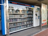 A vending machine filled with neatly arranged books is situated outdoors. There is a digital display above the machine showing text in a language that appears to be Chinese. Next to the machine is a panel with information and instructions, possibly related to how to operate it.