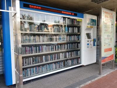 A vending machine filled with neatly arranged books is situated outdoors. There is a digital display above the machine showing text in a language that appears to be Chinese. Next to the machine is a panel with information and instructions, possibly related to how to operate it.