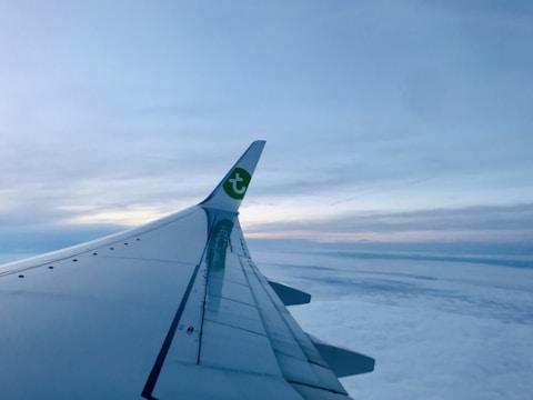 An airplane wing with a logo visible on the winglet extends across a cloud-covered sky during what appears to be sunset or sunrise, with blue and pink hues in the sky.