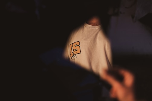 A white t-shirt with a partially visible bold orange and black graphic design is in the dimly lit scene. The focus is softly illuminated while the foreground remains shadowy with a hand holding a piece of black paper or object catching some light.