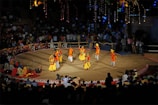 A traditional dance performance takes place on a circular stage surrounded by an audience. The dancers wear colorful, vibrant costumes with shades of yellow, orange, and red. The stage is outdoors, with decorative lights illuminating the background, and spectators watch from all sides.