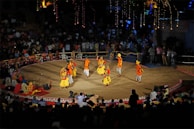 A traditional dance performance takes place on a circular stage surrounded by an audience. The dancers wear colorful, vibrant costumes with shades of yellow, orange, and red. The stage is outdoors, with decorative lights illuminating the background, and spectators watch from all sides.