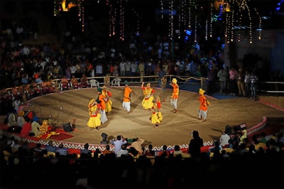 A traditional dance performance takes place on a circular stage surrounded by an audience. The dancers wear colorful, vibrant costumes with shades of yellow, orange, and red. The stage is outdoors, with decorative lights illuminating the background, and spectators watch from all sides.