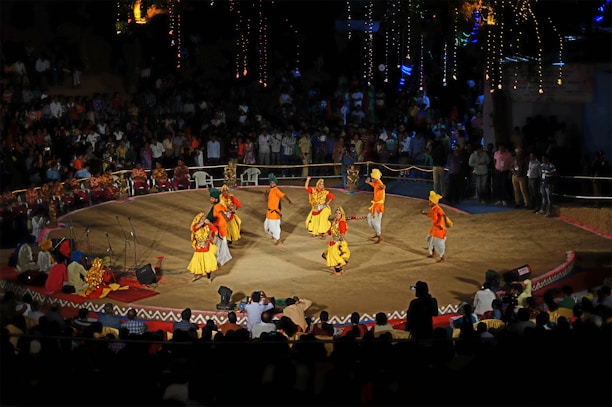 A traditional dance performance takes place on a circular stage surrounded by an audience. The dancers wear colorful, vibrant costumes with shades of yellow, orange, and red. The stage is outdoors, with decorative lights illuminating the background, and spectators watch from all sides.