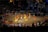 A traditional dance performance takes place on a circular stage surrounded by an audience. The dancers wear colorful, vibrant costumes with shades of yellow, orange, and red. The stage is outdoors, with decorative lights illuminating the background, and spectators watch from all sides.