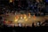 A traditional dance performance takes place on a circular stage surrounded by an audience. The dancers wear colorful, vibrant costumes with shades of yellow, orange, and red. The stage is outdoors, with decorative lights illuminating the background, and spectators watch from all sides.