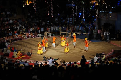 A traditional dance performance takes place on a circular stage surrounded by an audience. The dancers wear colorful, vibrant costumes with shades of yellow, orange, and red. The stage is outdoors, with decorative lights illuminating the background, and spectators watch from all sides.