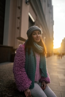 A person wearing a cozy outfit consisting of a pink fluffy jacket, teal scarf, and gray beanie. The setting sun casts a warm glow on the street lined with tall buildings.