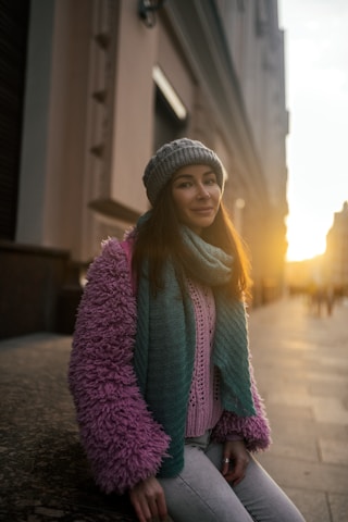 A person wearing a cozy outfit consisting of a pink fluffy jacket, teal scarf, and gray beanie. The setting sun casts a warm glow on the street lined with tall buildings.