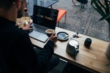 A person is sitting at a wooden table with a laptop displaying code on the screen. They are holding a cup of coffee. Nearby are a saucer with a spoon, a teapot, a camera lens, keys, and a glass bottle. The table is set beside a window providing an outside view of cobblestone pavement and part of a bicycle. There are plants partially visible, contributing to a cozy atmosphere.