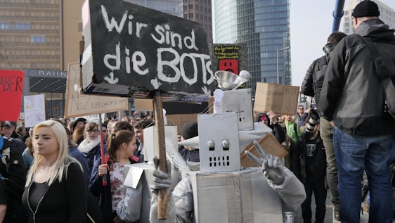 A crowd of people, some holding signs, are participating in a protest. Among them is a person dressed in a robot costume with a sign that reads 'Wir sind die Bots.' The atmosphere is busy and filled with various signs with messages related to digital rights.