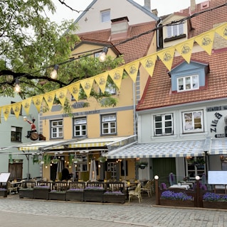 A charming street-side restaurant with colorful facades, featuring outdoor seating under striped awnings. Yellow bunting is strung across, with decorative lights adding brightness. Hanging flower baskets and well-tended planter boxes contribute a cozy, inviting atmosphere.