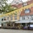 A charming street-side restaurant with colorful facades, featuring outdoor seating under striped awnings. Yellow bunting is strung across, with decorative lights adding brightness. Hanging flower baskets and well-tended planter boxes contribute a cozy, inviting atmosphere.