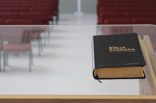 A black leather-bound Bible with gold lettering is placed on a transparent lectern or podium. In the background are rows of empty red chairs, suggesting a church or auditorium setting.