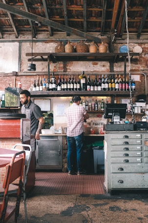 A rustic bar interior with exposed brick walls and wooden ceiling beams can be seen. Bottles of wine and spirits are arranged neatly on shelves, with wicker baskets placed on the higher shelf. Two individuals are present; one is wearing a checkered shirt and working behind the bar, while the other is approaching or serving. The setting suggests a casual and cozy atmosphere.