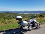 A modern motorcycle is parked on a roadside with a scenic view of a vast landscape in the background. The landscape includes lush green fields and hills extending into the distance. A body of water and a city are visible further away under a clear blue sky with scattered clouds.