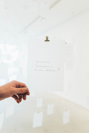A hand is holding a white piece of paper with handwritten text in a minimalist, bright setting. The room has a soft focus with several similar papers hanging on strings in the background.