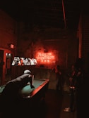 A dimly lit bar scene with a neon sign on the wall that reads 'Anejo Pendejo'. A pool table is in the center with a man leaning over, aiming a pool cue. Three people are observing from the side. The lighting is predominantly red and the ambiance appears rustic and edgy.