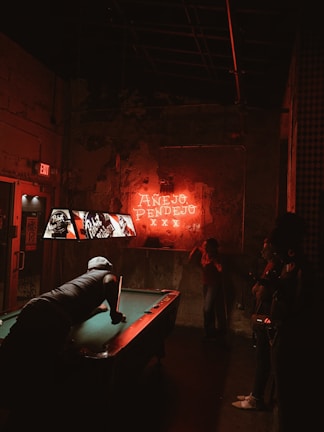 A dimly lit bar scene with a neon sign on the wall that reads 'Anejo Pendejo'. A pool table is in the center with a man leaning over, aiming a pool cue. Three people are observing from the side. The lighting is predominantly red and the ambiance appears rustic and edgy.