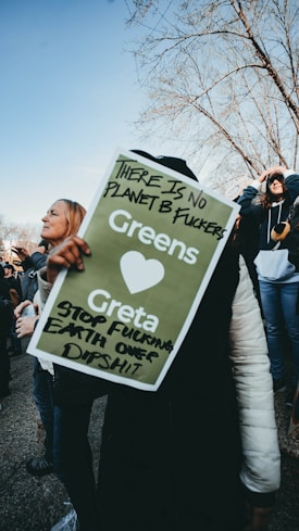 A group of people outdoors holding a protest sign with messages about climate change. The sign is green with white text and some black handwritten notes. The weather appears clear, and some trees without leaves are visible in the background.