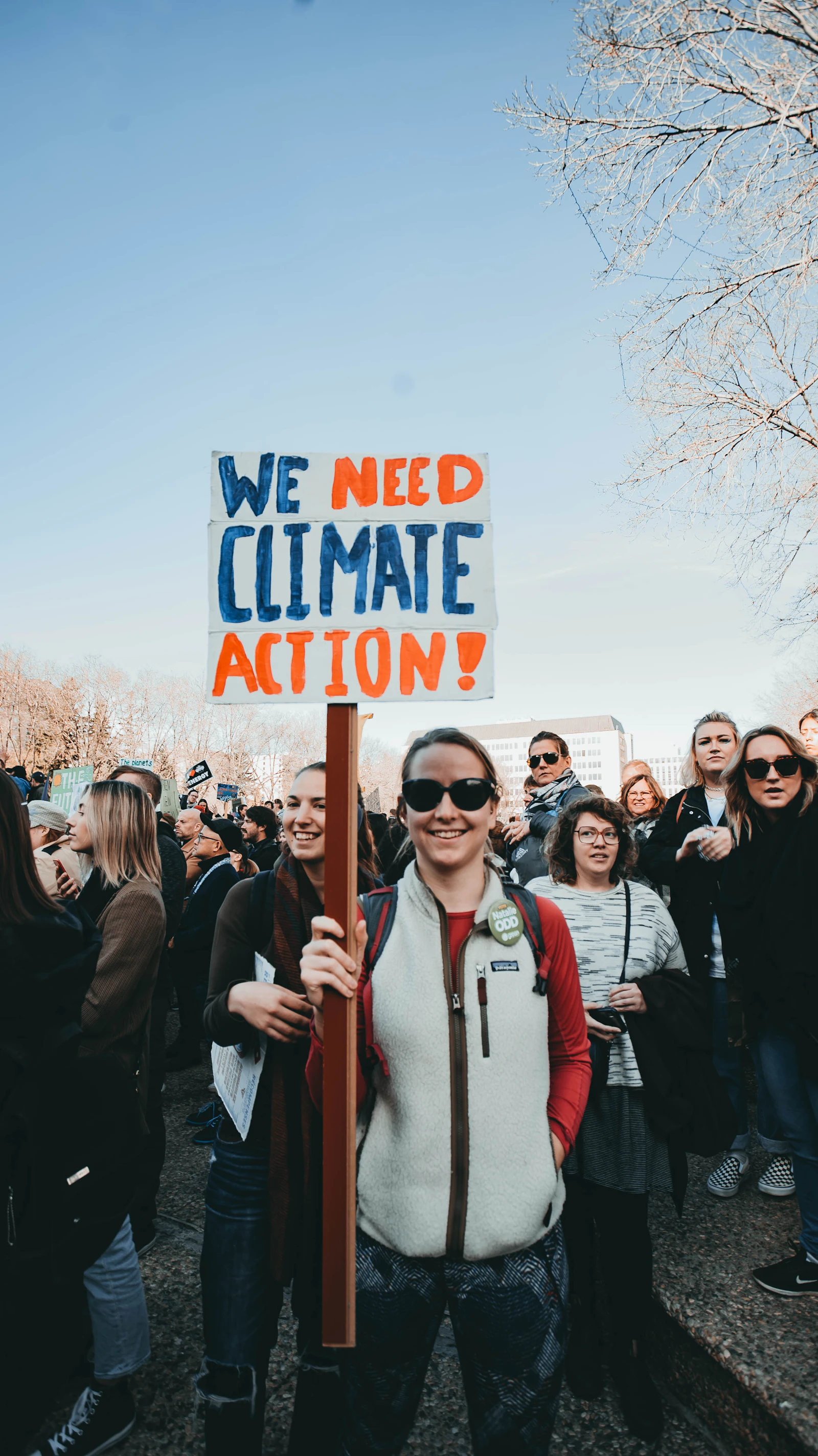 A woman standing outdoors, smiling while holding a sign that reads 'We Need Climate Action!'