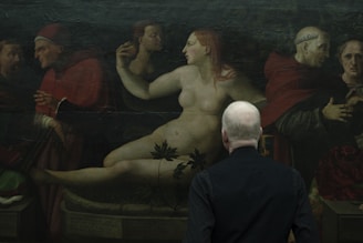 A person with gray hair, seen from behind, is observing an artwork depicting a group of historical figures, including a nude woman reclining among clothed figures wearing robes and head coverings. The setting appears dark, conveying a classical and historical atmosphere.