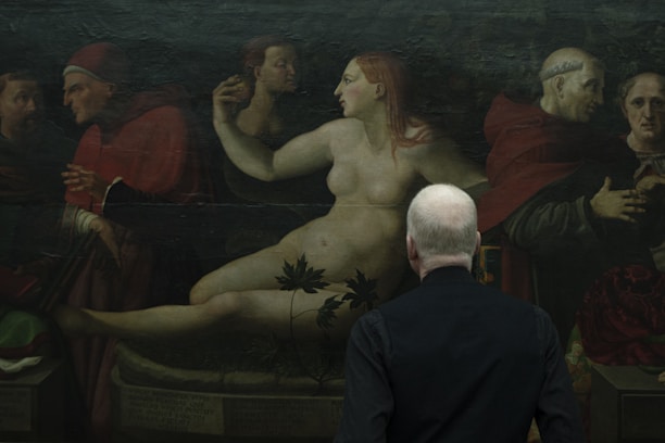 A person with gray hair, seen from behind, is observing an artwork depicting a group of historical figures, including a nude woman reclining among clothed figures wearing robes and head coverings. The setting appears dark, conveying a classical and historical atmosphere.