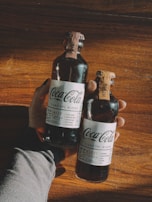 A hand is holding two glass bottles of Coca-Cola Signature Mixers placed on a wooden surface. The bottles have a dark liquid inside and feature labels indicating different flavors, such as 'Smoky' and 'Woody'. The warm sunlight creates a cozy atmosphere, casting shadows across the wood.