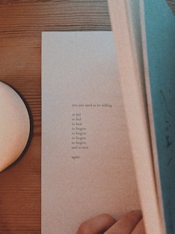 A person is holding open a book on a wooden table. The page contains inspirational text that speaks about being willing to fail, feel, heal, forgive, and love again.