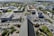 The image provides an aerial view of a cityscape with a prominent triangular architectural structure in the foreground, surrounded by parked cars and green landscaping. Various rectangular buildings and streets extend to the horizon, with patches of greenery interspersed throughout the urban area.