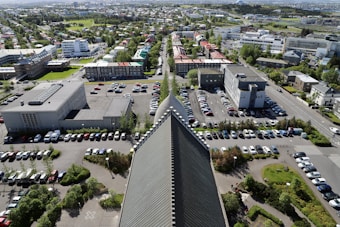 The image provides an aerial view of a cityscape with a prominent triangular architectural structure in the foreground, surrounded by parked cars and green landscaping. Various rectangular buildings and streets extend to the horizon, with patches of greenery interspersed throughout the urban area.