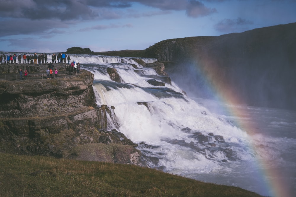 photography of people standing while watching waterfalls covered by rainbow