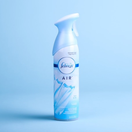 A Febreze air freshener spray can with a light blue and white design is positioned against a matching light blue background. The can features an image of flowing fabric and the words 'AIR' along with the scent 'linen & sky'.