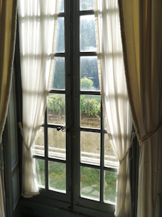 Sunlight filters through sheer white curtains on a tall, grid-paneled window that looks out onto a garden with lush greenery. The interior features soft, gently pulled-back drapery framing the window, creating a serene and inviting atmosphere.