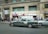 A busy urban street scene with a green sedan showing signs of wear, parked in front of a building with the inscription 'Pasaje Catedral'. Several people are walking or standing nearby, with some gathered near a storefront with signs advertising currency exchange and other services. The building exterior is concrete with windows on the upper floors, and there are various signs and advertisements visible.