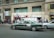 A busy urban street scene with a green sedan showing signs of wear, parked in front of a building with the inscription 'Pasaje Catedral'. Several people are walking or standing nearby, with some gathered near a storefront with signs advertising currency exchange and other services. The building exterior is concrete with windows on the upper floors, and there are various signs and advertisements visible.