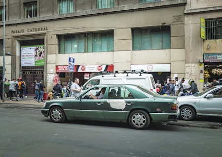A busy urban street scene with a green sedan showing signs of wear, parked in front of a building with the inscription 'Pasaje Catedral'. Several people are walking or standing nearby, with some gathered near a storefront with signs advertising currency exchange and other services. The building exterior is concrete with windows on the upper floors, and there are various signs and advertisements visible.