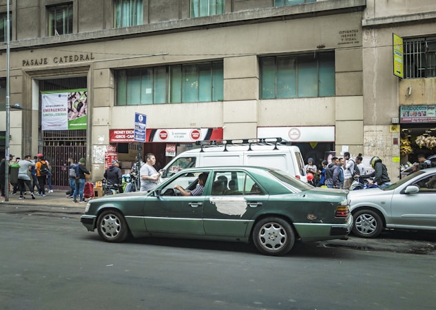 A busy urban street scene with a green sedan showing signs of wear, parked in front of a building with the inscription 'Pasaje Catedral'. Several people are walking or standing nearby, with some gathered near a storefront with signs advertising currency exchange and other services. The building exterior is concrete with windows on the upper floors, and there are various signs and advertisements visible.