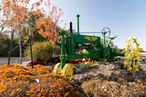 A bright green John Deere tractor is prominently featured in a landscaped garden area surrounded by blooming flowers and autumnal trees with orange leaves. The vibrant tractor stands on a mound of multicolored flowers and mulch, adding to the picturesque garden setting.