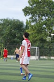 A soccer player wearing a red and white jersey with the number 23 is walking on a green field. In the background, other players are visible, including one without a shirt. There are trees and a goalpost in the distance under a clear sky.