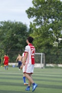 A soccer player wearing a red and white jersey with the number 23 is walking on a green field. In the background, other players are visible, including one without a shirt. There are trees and a goalpost in the distance under a clear sky.