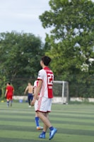 A soccer player wearing a red and white jersey with the number 23 is walking on a green field. In the background, other players are visible, including one without a shirt. There are trees and a goalpost in the distance under a clear sky.