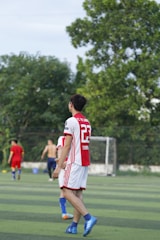 A soccer player wearing a red and white jersey with the number 23 is walking on a green field. In the background, other players are visible, including one without a shirt. There are trees and a goalpost in the distance under a clear sky.