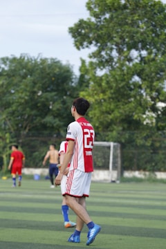 A soccer player wearing a red and white jersey with the number 23 is walking on a green field. In the background, other players are visible, including one without a shirt. There are trees and a goalpost in the distance under a clear sky.