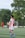 A soccer player wearing a red and white jersey with the number 23 is walking on a green field. In the background, other players are visible, including one without a shirt. There are trees and a goalpost in the distance under a clear sky.