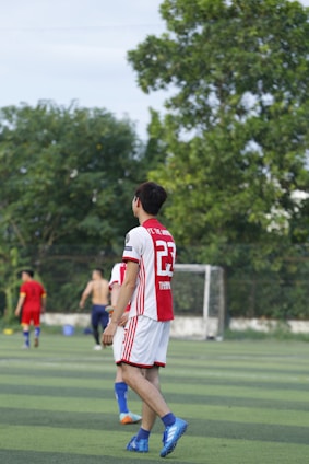 A soccer player wearing a red and white jersey with the number 23 is walking on a green field. In the background, other players are visible, including one without a shirt. There are trees and a goalpost in the distance under a clear sky.