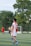 A soccer player wearing a red and white jersey with the number 23 is walking on a green field. In the background, other players are visible, including one without a shirt. There are trees and a goalpost in the distance under a clear sky.