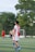 A soccer player wearing a red and white jersey with the number 23 is walking on a green field. In the background, other players are visible, including one without a shirt. There are trees and a goalpost in the distance under a clear sky.